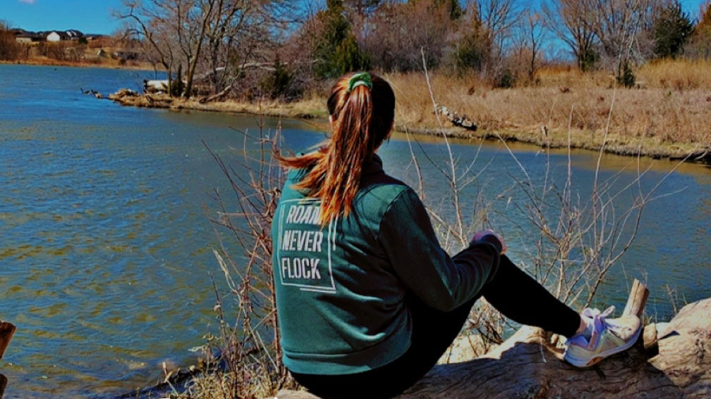 Woman in green jacket with 'Roam Never Flock' text sits by river in nature landscape