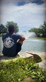 Person in LynXautoinnovate shirt sitting by pond with fountain, lush grass, blue sky