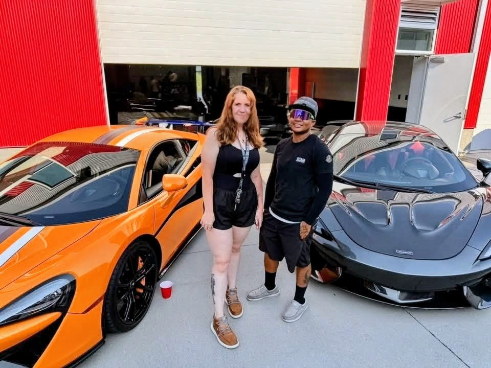 Two people standing outdoors between two sports cars in front of a garage building. The left car is bright orange with black racing stripes, and the right car is a dark gray McLaren.
